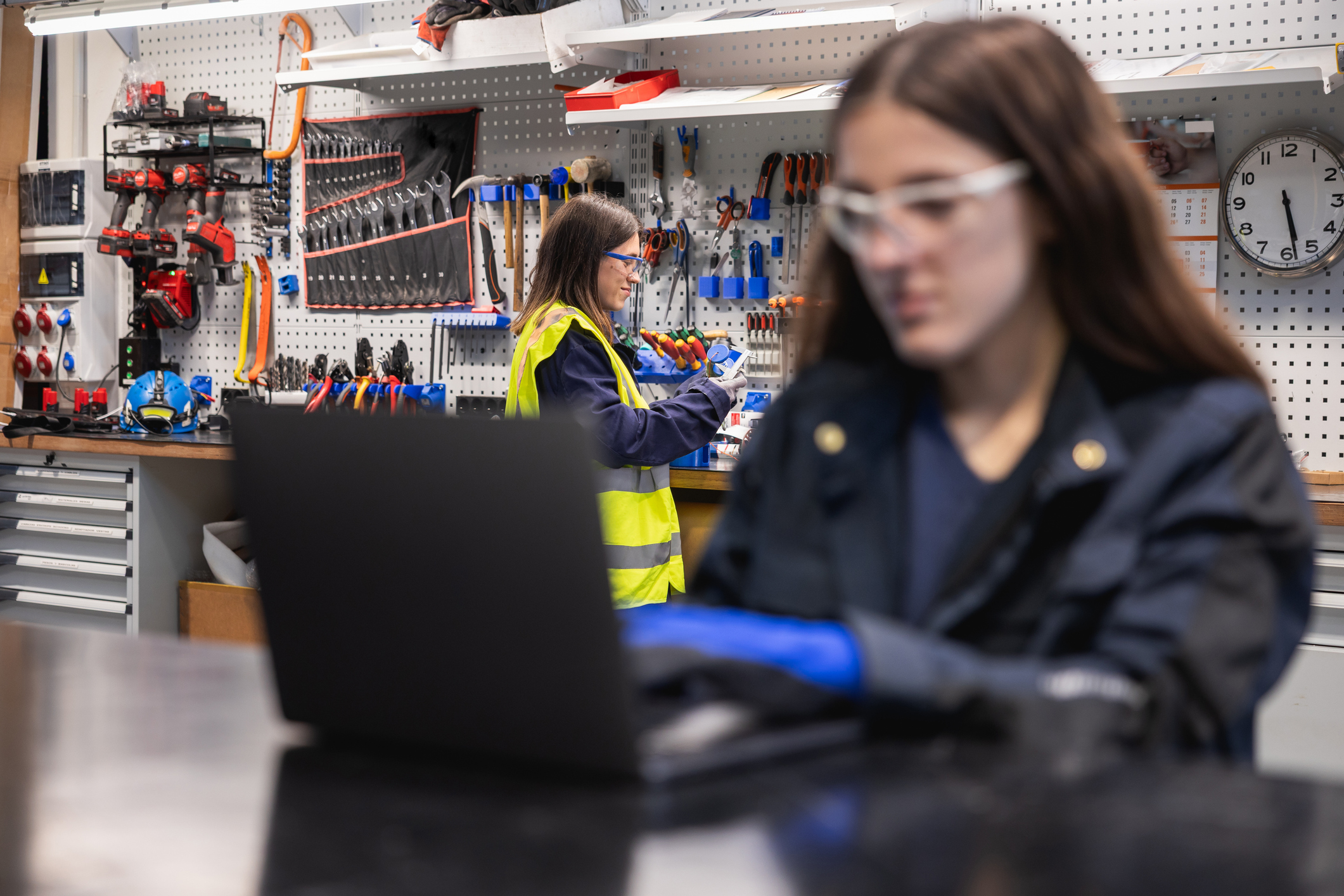 Young women working in a workshop, one using a laptop and another organizing tools.