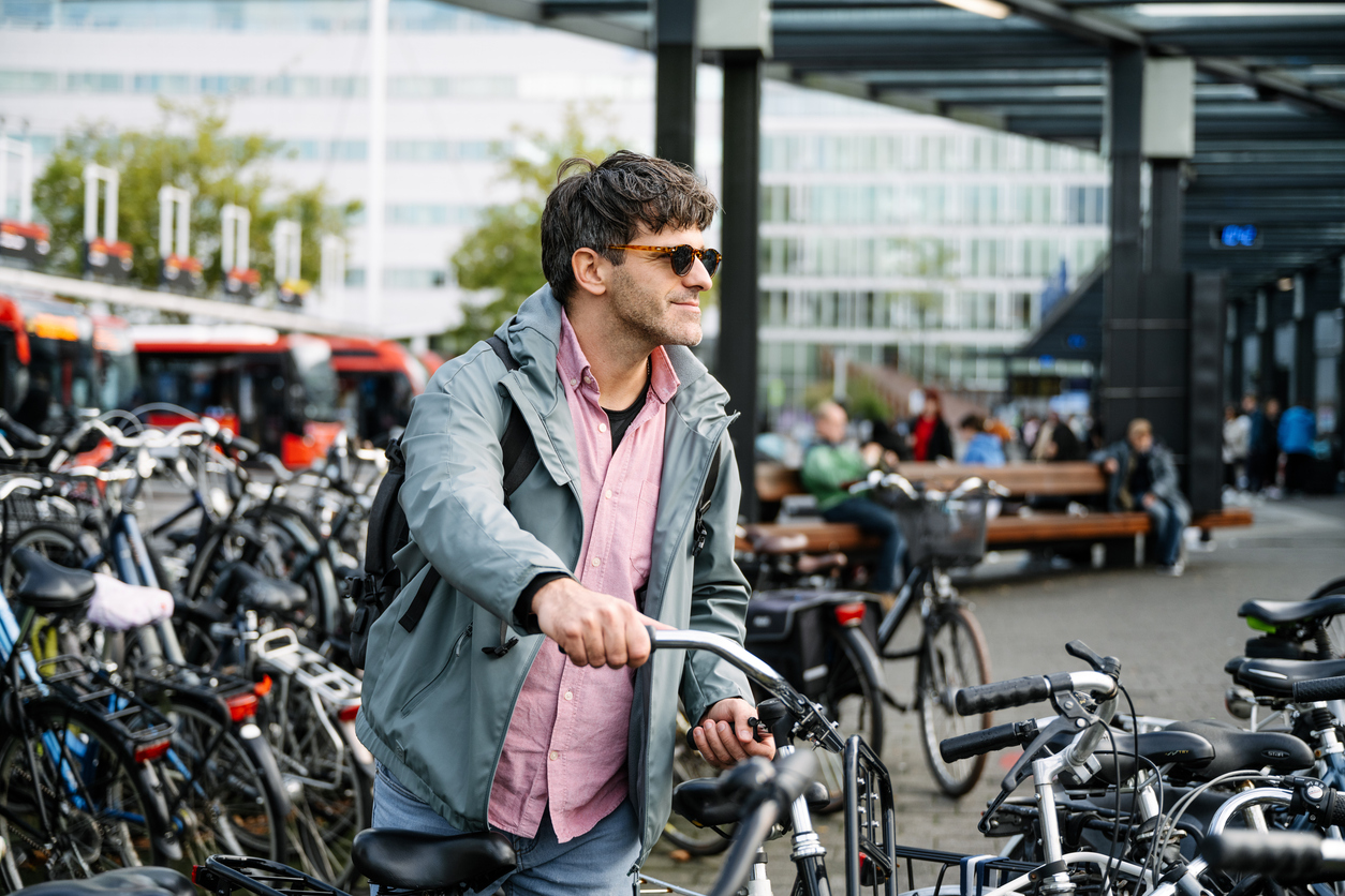 Adult man in casual attire stands with his bicycle at a busy urban transit station during the day, surrounded by parked bikes and public transport buses.